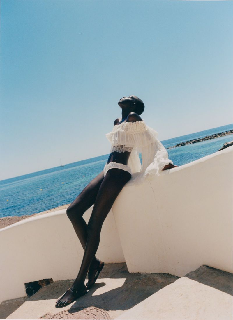 Model in white ruffled top and bikini bottoms leaning against a white wall by the ocean under clear sky