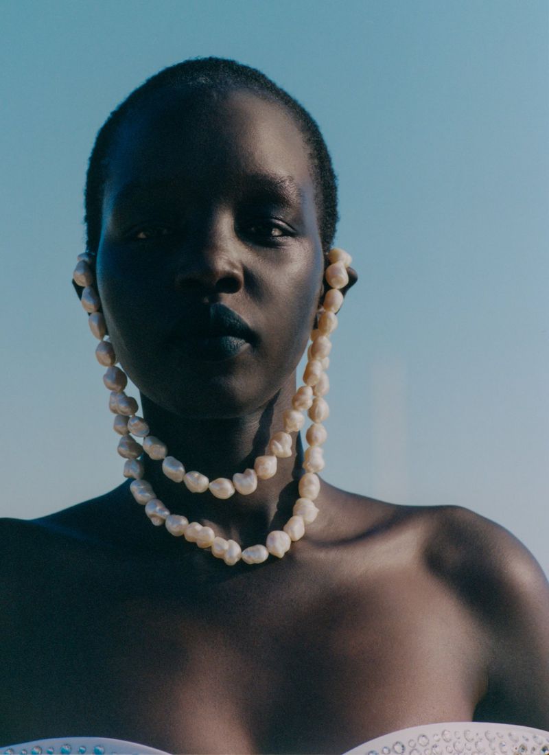 Model Akuol Deng Atem wearing a draped baroque pearl necklace connected to earrings against an open blue sky.