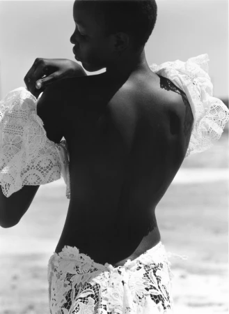 Akuol Deng Atem facing away in a white lace garment with a bare back on a sunlit beach.