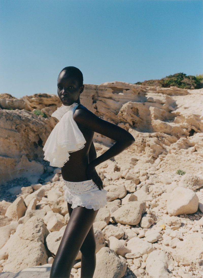 Model Akuol Deng Atem poses in white ruffled garments against a rugged rocky landscape under a blue sky.