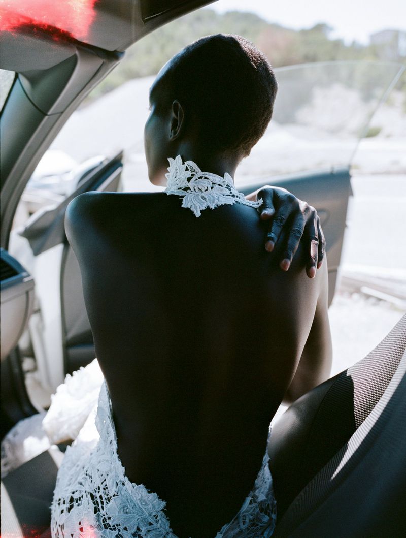 Model Akuol Deng Atem seated in a car with her back turned, wearing an open-back white floral lace garment.