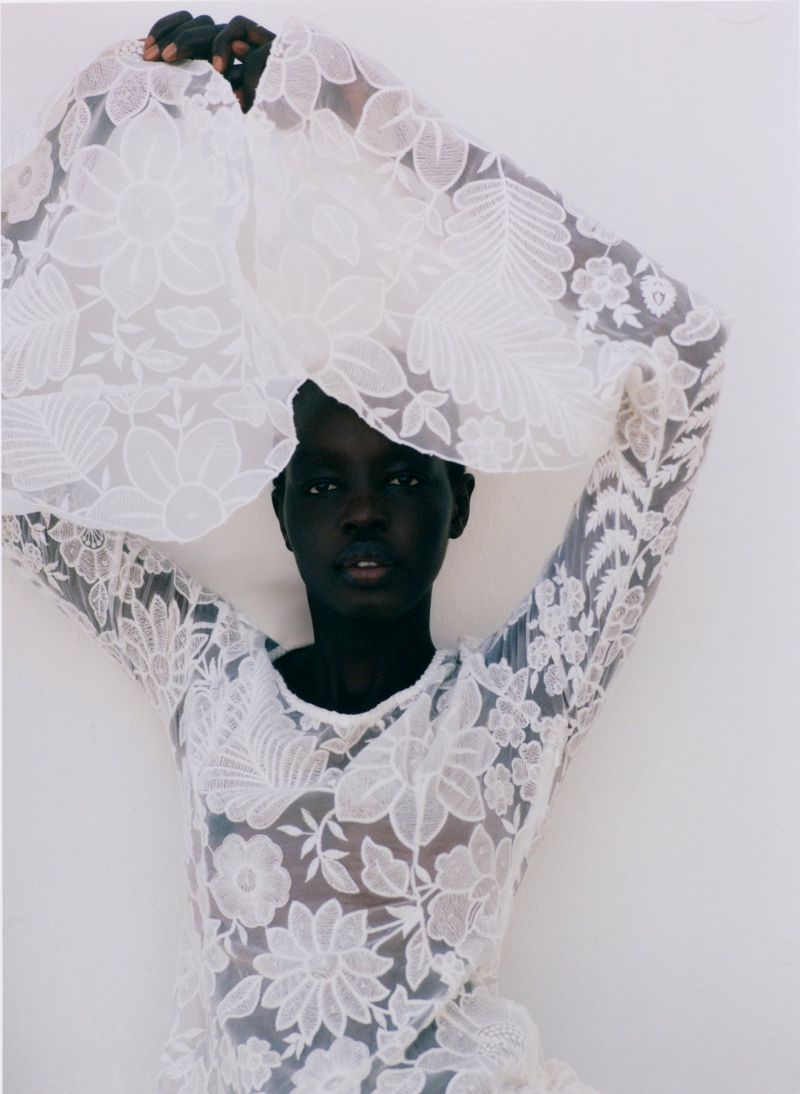 Model Akuol Deng Atem posing with raised arms in a sheer white floral lace dress against a plain white studio background.