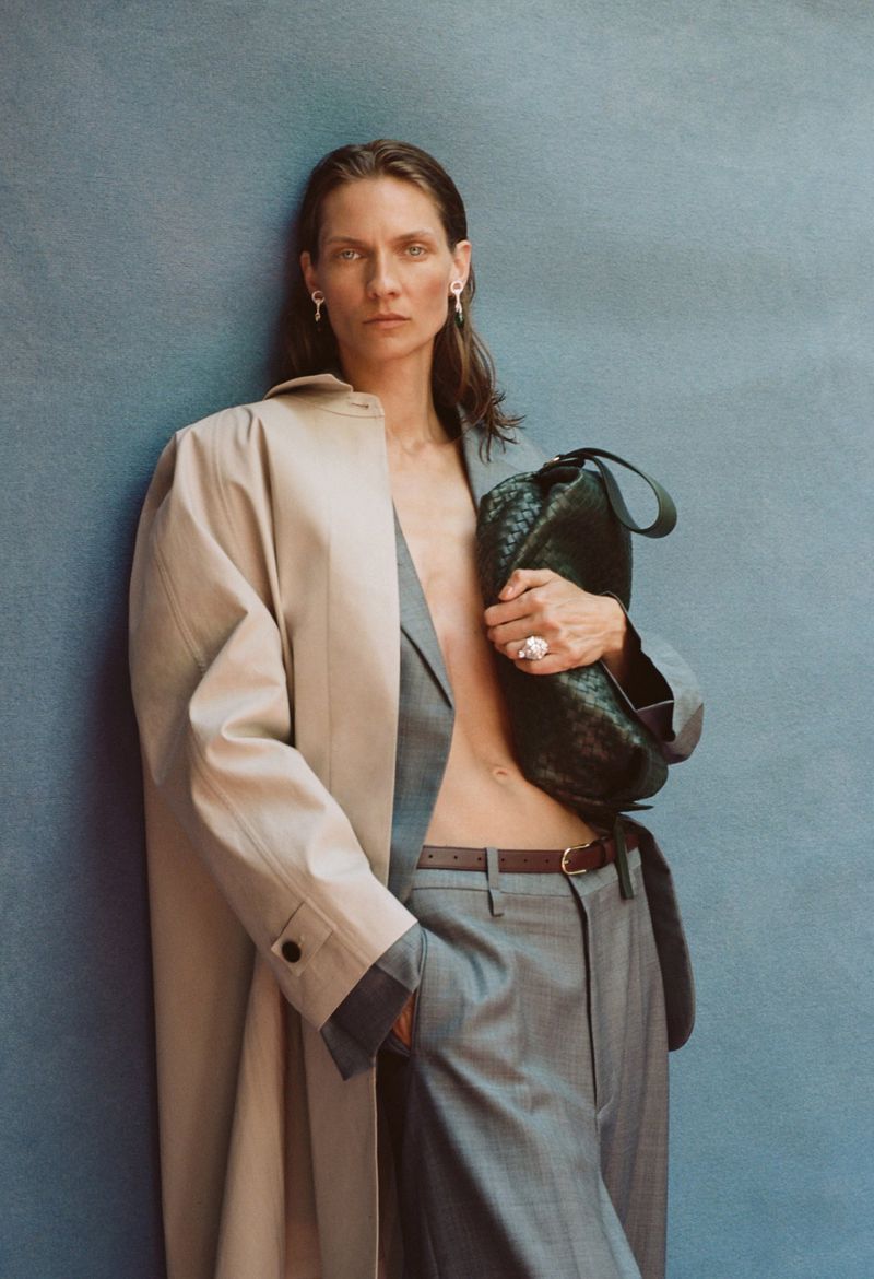 Karolin Wolter wearing a grey suit and beige trench coat holding a dark green woven handbag against a blue wall.