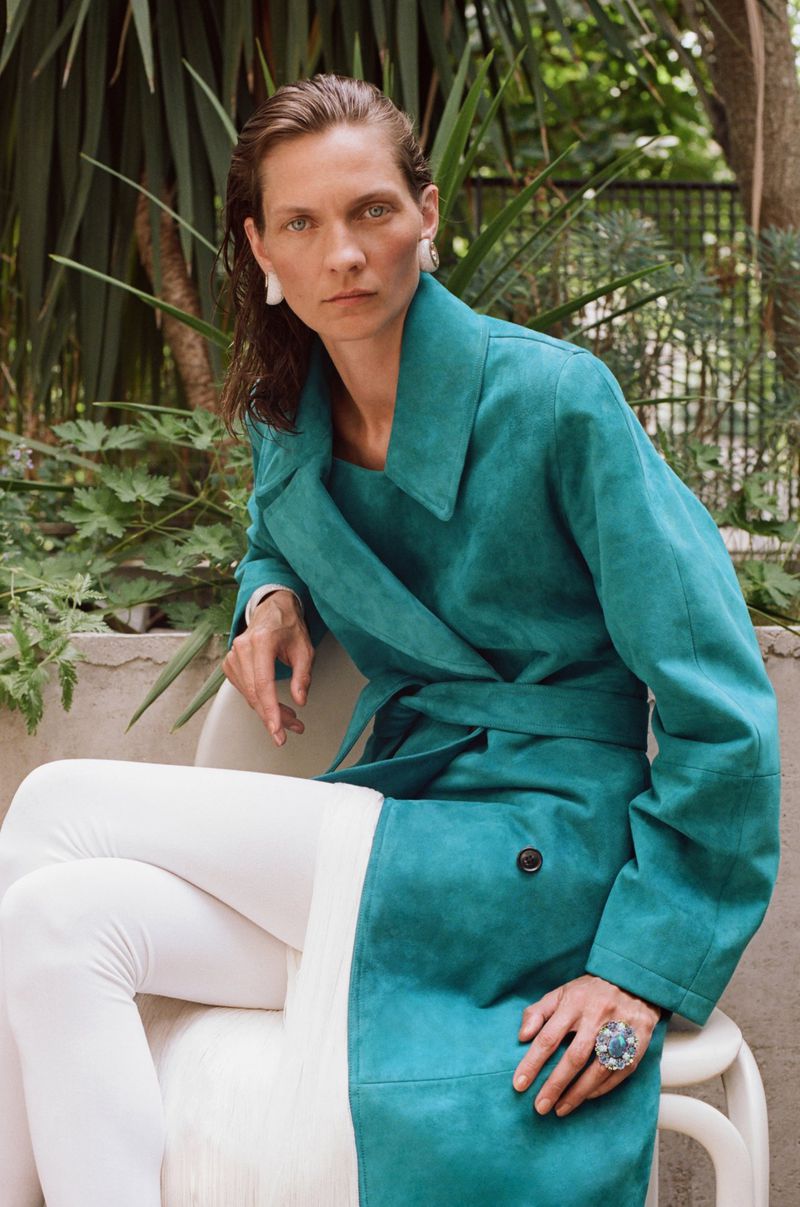 Model Karolin Wolter seated in a vivid teal belted trench coat and white tights in an outdoor garden setting.