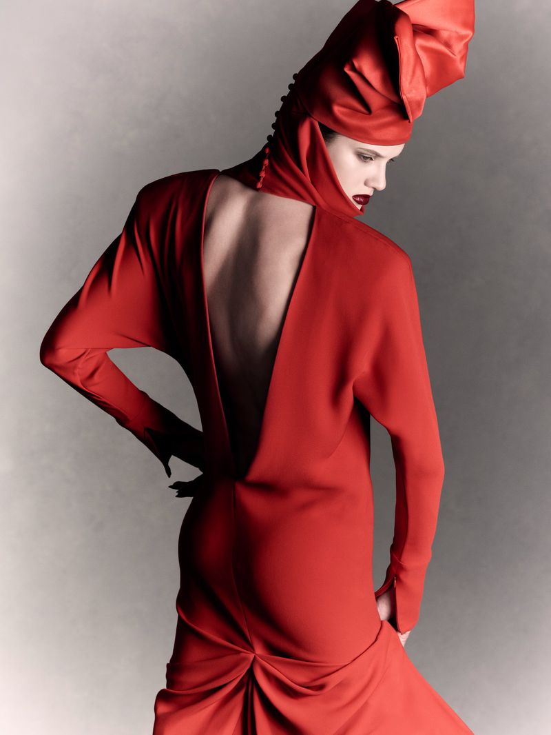 Model Noor Khan wearing a backless red gown with a sculpted hood against a grey backdrop for V Magazine.