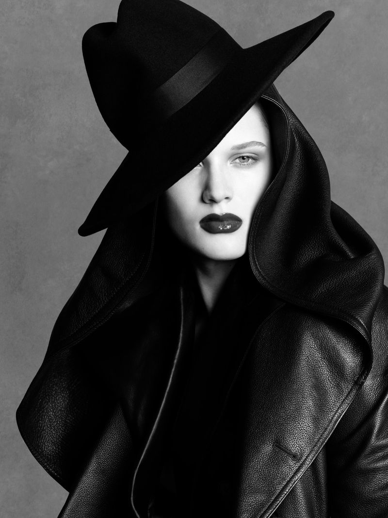 Monochrome close-up portrait of model Noor Khan wearing a black wide-brim hat and hooded leather coat against a grey studio backdrop.