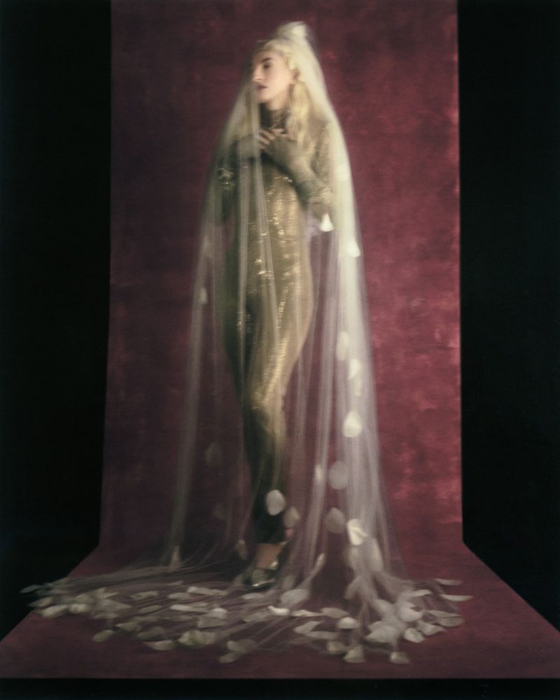 Model Riley Woodell standing in a shimmering gold jumpsuit beneath a floor-length sheer veil on a deep burgundy textured backdrop.