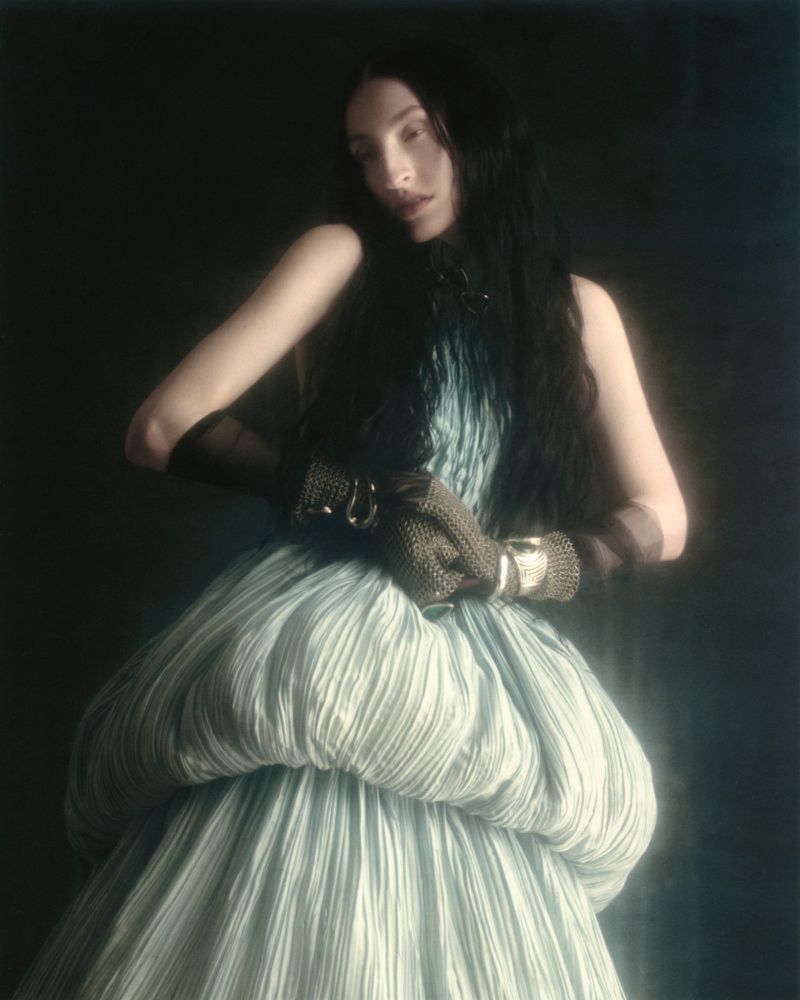Model Riley Woodell posing in a voluminous pleated pale blue gown with textured brown gloves against a dark backdrop.