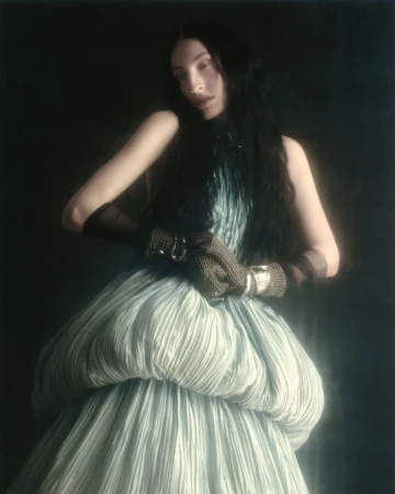Model Riley Woodell posing in a voluminous pleated pale blue gown with textured brown gloves against a dark backdrop.