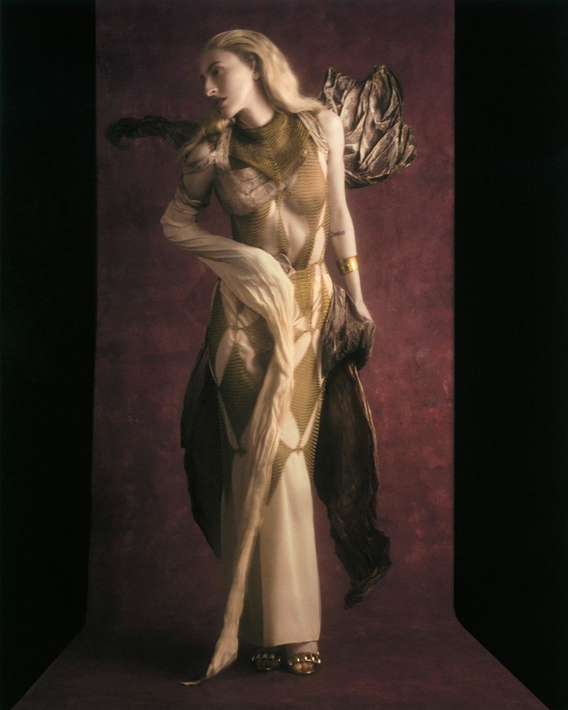 Model Riley Woodell poses in an intricate gold mesh and draped cream gown against a deep textured burgundy backdrop.