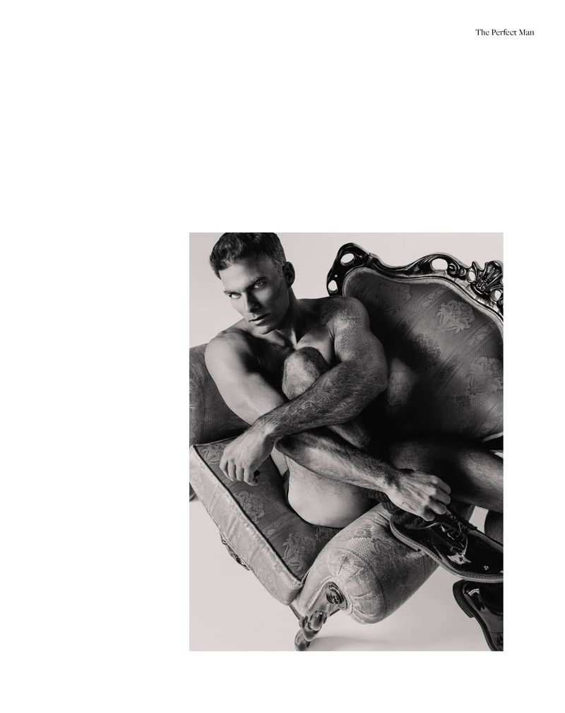 Monochrome editorial portrait of a shirtless man seated curled in an ornate vintage armchair wearing black patent shoes.