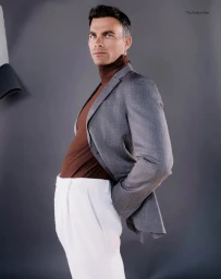 Man in a grey blazer, brown turtleneck, and white trousers posing against a grey studio backdrop.