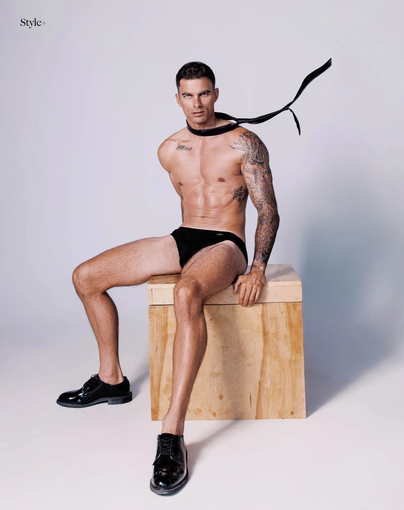 A shirtless man in black briefs and shoes sits on a wooden crate with a floating black tie against a white studio background.