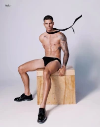 A shirtless man in black briefs and shoes sits on a wooden crate with a floating black tie against a white studio background.