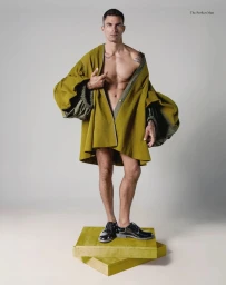 Man standing on yellow platform blocks wearing an open voluminous olive green cape and black dress shoes in a studio setting.