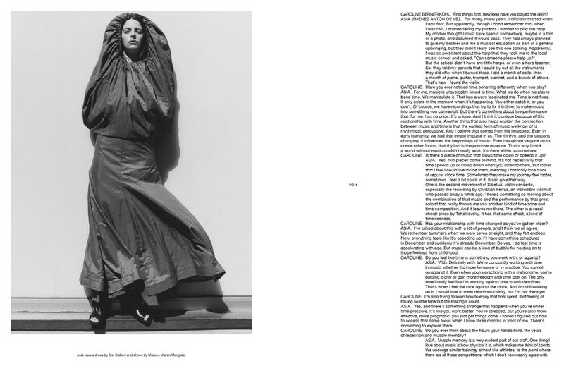 Monochromatic fashion portrait of a woman in an oversized hooded dress, standing against a plain wall in a studio-like setting.