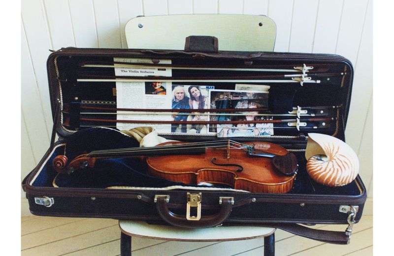 Open violin case with brown violin, music sheets, and shell resting inside, set on a chair against a white paneled wall.
