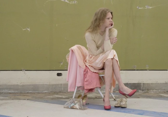 Model Wolfgang Beck seated on a chair wearing a cream top, pink skirt, and red heels in front of an olive green wall.