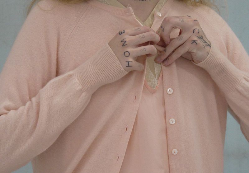 Close-up of a person with hand tattoos wearing a pale peach knit cardigan over a matching lace-trimmed top.