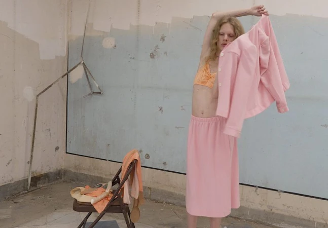 Model Wolfgang Beck posing in a pink skirt and orange bralette while holding up a matching pink jacket in a distressed room.