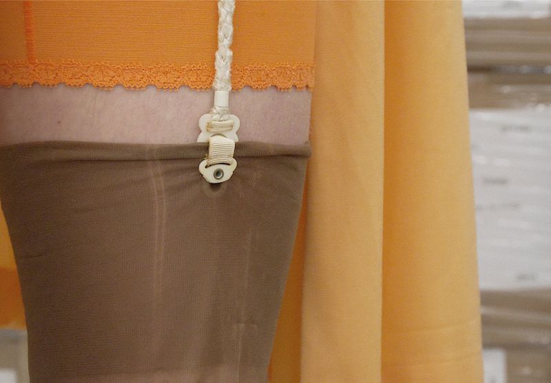Close-up detail of a white braided garter strap and clip attached to a sheer brown stocking and orange undergarment.