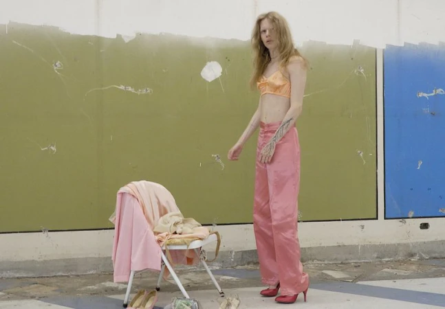 Model Wolfgang Beck stands in a peach satin bra top and pink satin trousers beside a folding chair with draped clothes.
