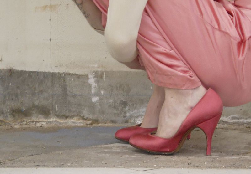 Close-up of a person crouching outdoors wearing pink satin high heels and matching pink draped silk trousers against a concrete wall.
