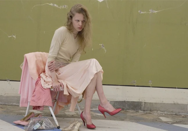 Model seated on folding chair wearing cream sweater and pink skirt against weathered green wall for ALL-IN editorial