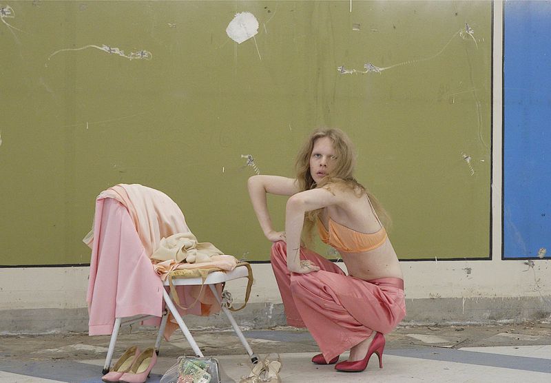 Model Wolfgang Beck crouching in pink satin trousers and an orange bralette beside a draped folding chair against an olive green wall.