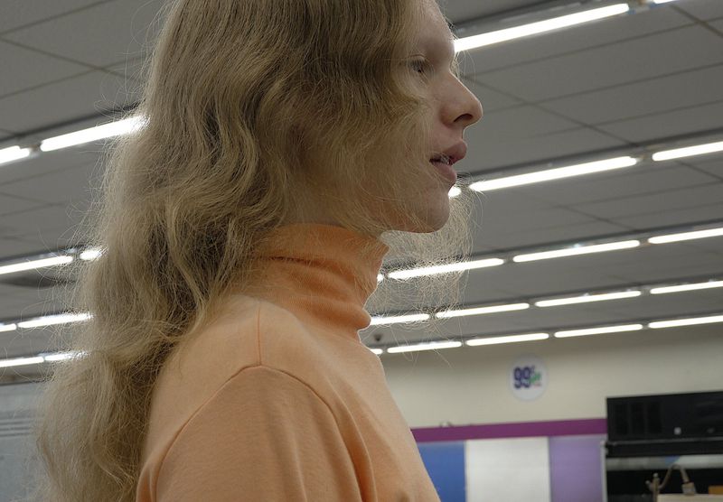 Profile portrait of a long-haired model wearing a peach turtleneck sweater inside a brightly lit retail store interior.