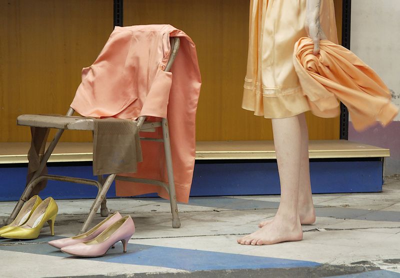 Barefoot person standing in a peach silk skirt beside a metal chair draped with garments and discarded high heels on a tiled floor.