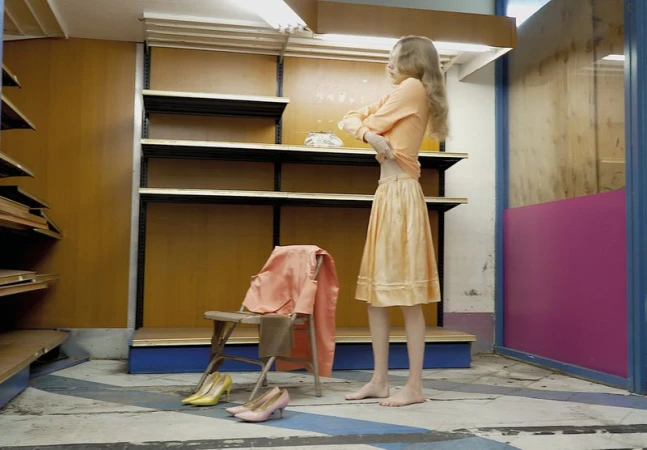 Model Wolfgang Beck stands barefoot in a peach skirt and top beside empty retail shelving and heels on the floor.