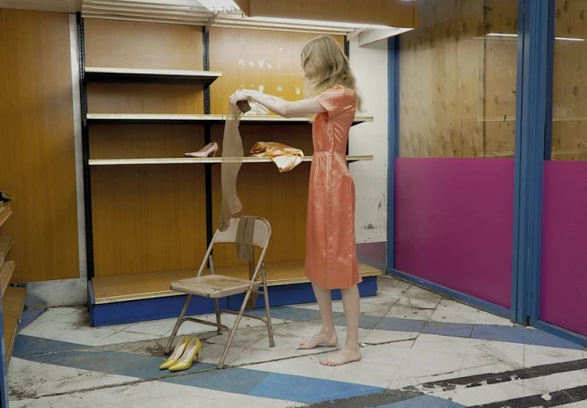 A barefoot person in a peach satin dress stands holding pantyhose beside a folding chair and yellow heels in an empty store.