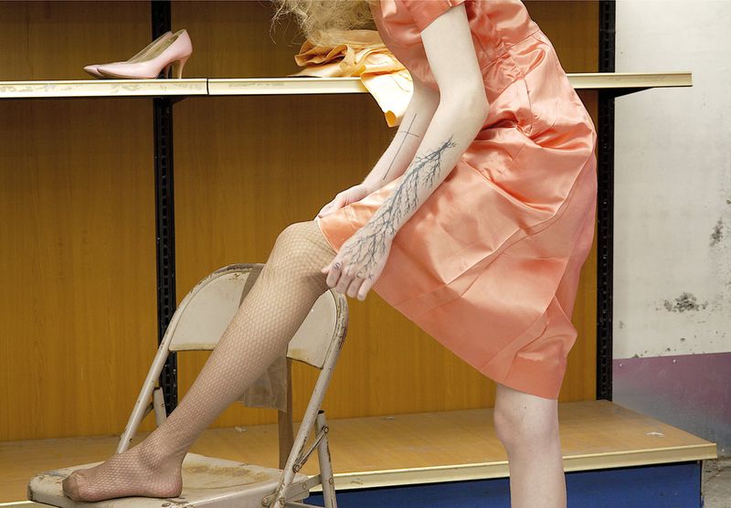 Model in a peach satin dress placing one leg on a folding chair, displaying patterned mesh tights and arm tattoos.