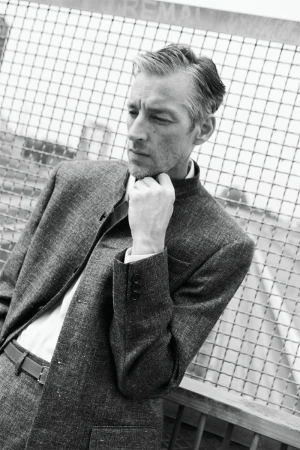 Model Joel Frampton in a textured grey stand-collar tailored suit posing with hand on chin against a wire mesh fence in black and white.