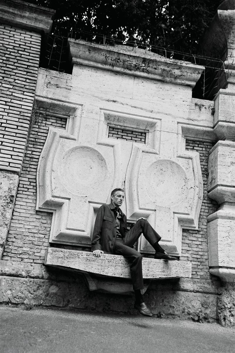 Man seated on an ornate stone wall outdoors in a monochrome editorial fashion photograph.