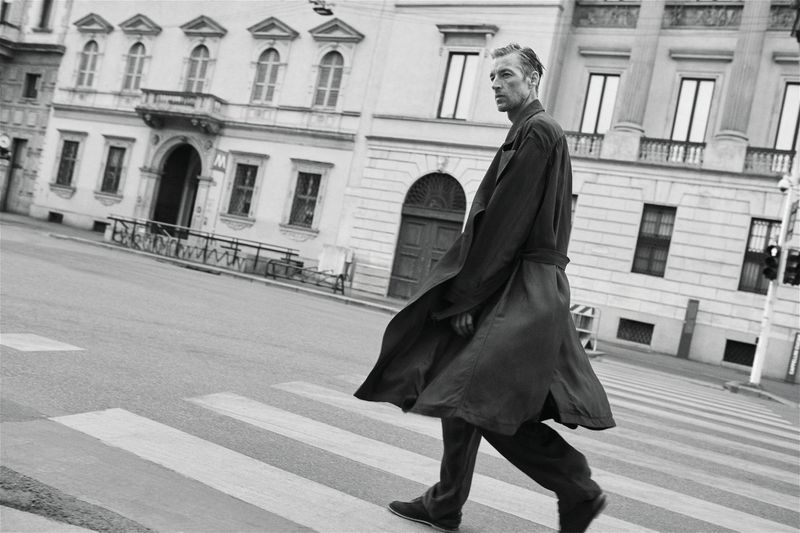 Joel Frampton wearing a long dark trench coat while walking across a crosswalk on a city street in front of classical architecture.