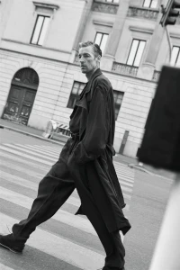 Model Joel Frampton walks across a crosswalk wearing an oversized dark trench coat and wide-leg trousers in a city street.
