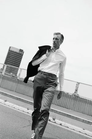 Model Joel Frampton walking in a white shirt and trousers with a jacket slung over his shoulder against an urban backdrop.