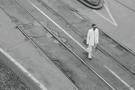 High-angle black and white shot of a man wearing a white suit and sunglasses walking across tram tracks on an asphalt road.