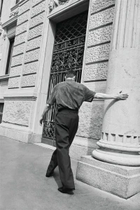 Black and white photo of model Joel Frampton walking from behind along a building exterior with his hand resting against a stone column.