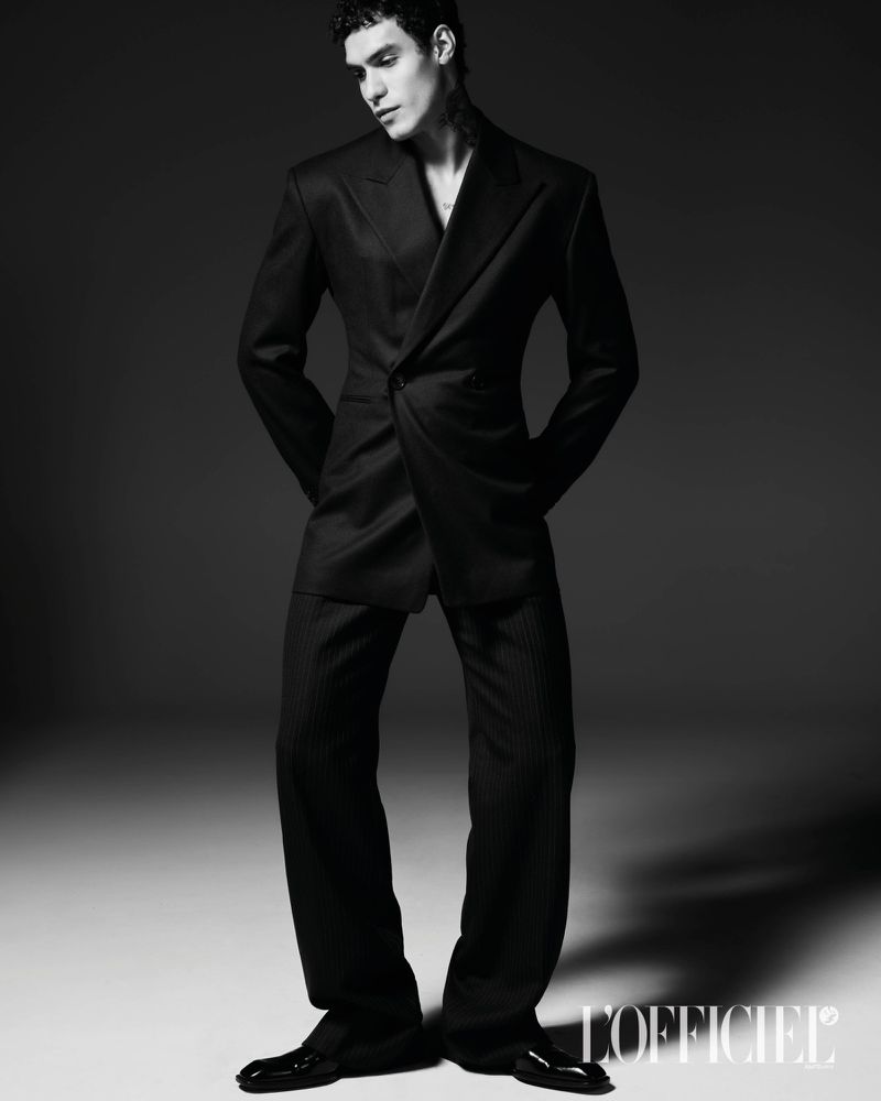 Model Leandro da Silva poses in a tailored black suit and dress shoes against a dark studio background in black and white.