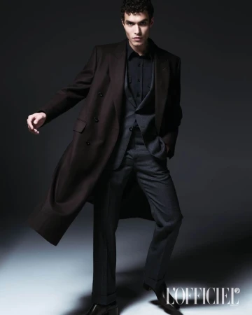 Model Leandro da Silva wearing a dark tailored overcoat and suit posing against a moody dark studio background.