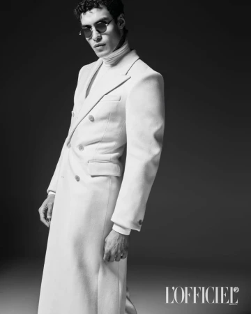 Model Leandro da Silva wearing a white double-breasted overcoat and dark sunglasses against a dark studio background.