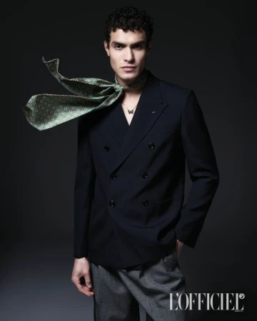 Model Leandro da Silva in a double-breasted navy blazer with a windswept green patterned silk scarf against a dark background.