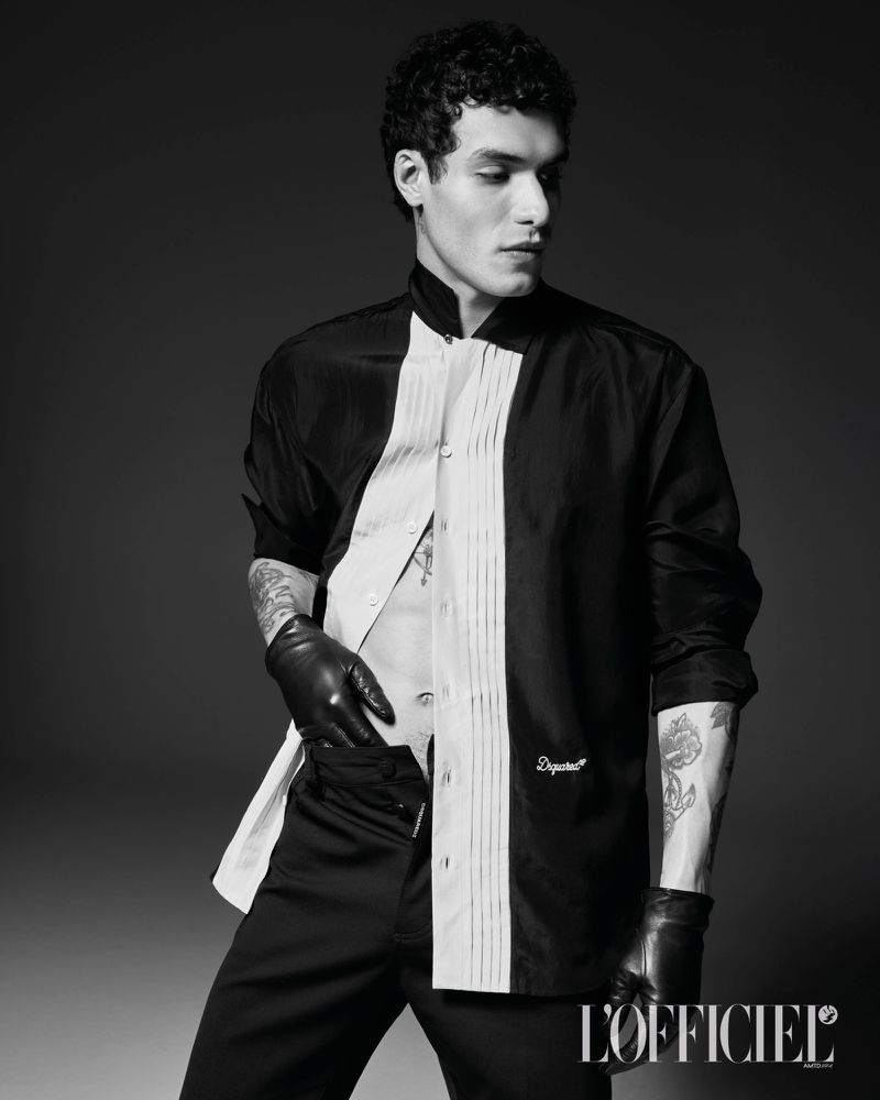 Model Leandro da Silva posing in an open black-and-white pleated shirt and black leather gloves against a dark studio backdrop.