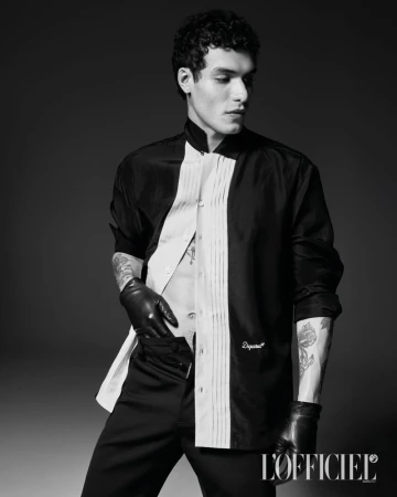 Model Leandro da Silva posing in an open black-and-white pleated shirt and black leather gloves against a dark studio backdrop.