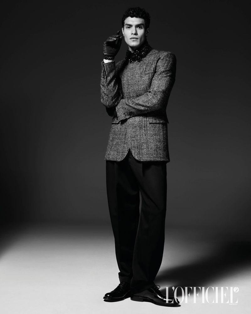 Model Leandro da Silva wearing a textured blazer, black trousers, and gloves in a monochrome studio portrait.
