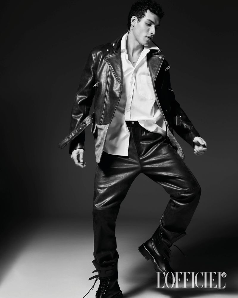 Model Leandro da Silva posing dynamically in a leather jacket, white dress shirt, and black trousers against a dark backdrop.