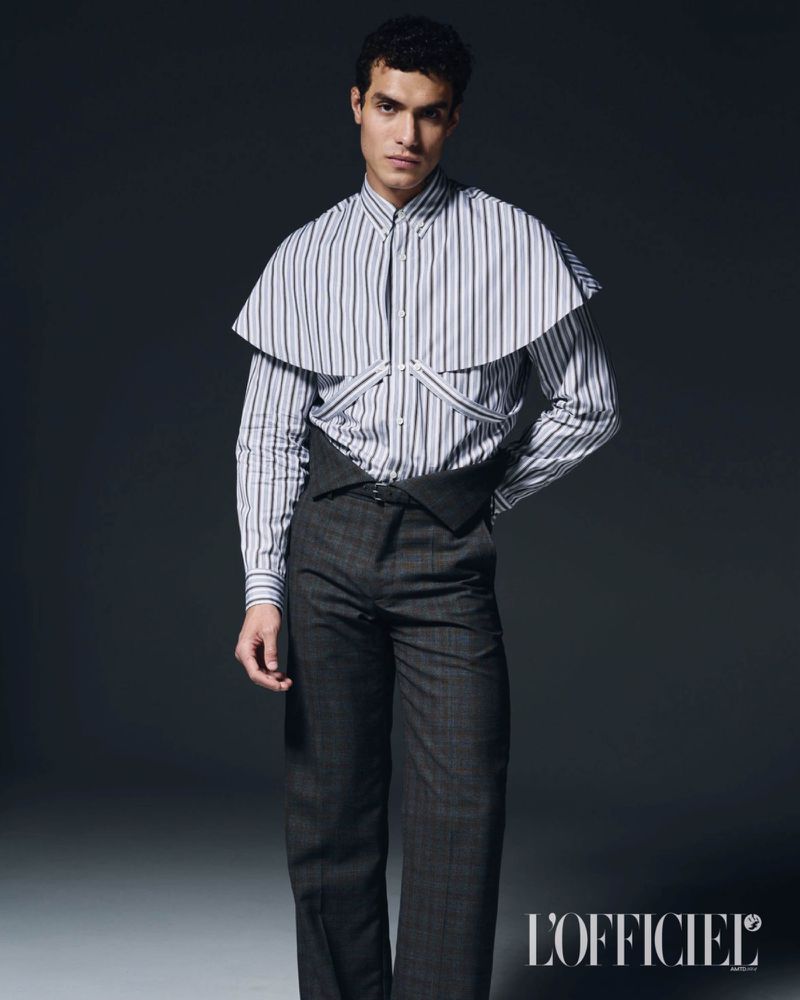 Model Leandro da Silva wearing a striped shirt with capelet overlay and high-waisted dark trousers against a dark studio background.